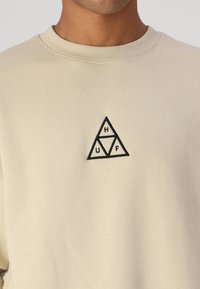 Felpa beige con scollatura rotonda e tessuto testurizzato. Include un piccolo logo triangolare ricamato in nero con le lettere "H", "U" e "F" all'interno.