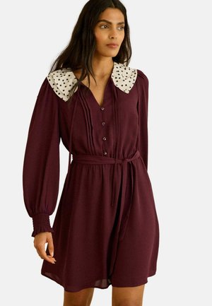 Moteris apsirengusi ilgomis rankovėmis violetinės spalvos suknele su diržu ties juosmeniu ir balta apykakle su juodais taškeliais, stovinti priešais vienspalvį foną.