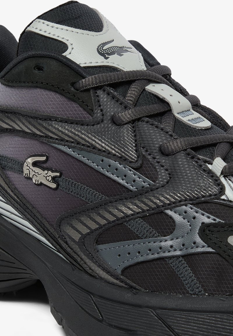 Schwarzer und grauer Lacoste-Sneaker mit strukturierten Paneelen, Schnürsenkeln und einem Krokodil-Logo an der Seite und auf der Zunge.