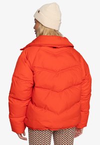 Giacca a vento arancione con motivo a zig-zag, colletto ampio e polsini elasticizzati. Indossata con pantaloni patterned e un berretto in maglia crema.