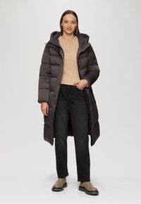 Manteau rembourré long de couleur gris foncé avec une capuche, doté d'une fermeture éclair et de coutures horizontales, accompagné d'un pull en tricot léger et d'un jean noir.