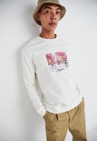 White sweatshirt with a pink graphic of a street scene, featuring the text "New York" and a khaki hat, paired with beige trousers.