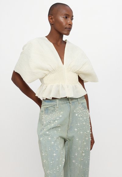 Hvid struktureret bluse med dyb V-udskæring og peplum-nederdel, parret med lyseblå jeans prydet med hvide perler på de forreste lommer.