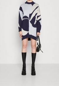 Abito lavorato a maglia con collo alto, caratterizzato da motivi astratti in blu navy, azzurro chiaro e crema. Abbinato a stivali neri al ginocchio.