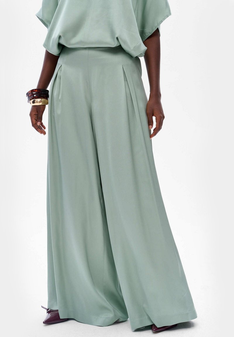 Femme portant un pantalon ample vert clair à jambes larges, assorti à un haut coordonné, accessoirisée avec des bracelets et des chaussures pointues violettes.