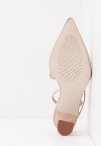 Semelle d'une chaussure à talon haut beige à bout pointu avec une semelle texturée et un talon bloc, positionnée à la verticale contre un mur d'angle blanc.