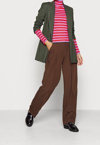 Kvinna klädd i en olivgrön kavaj, lila och rödrandig polotröja, bruna vida byxor och svarta loafers, stående med en hand i fickan.