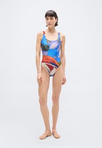 Γυναίκα που στέκεται ξυπόλητη φορώντας πολύχρωμο ολόσωμο μαγιό με λουλούδια, μπροστά σε ουδέτερο ανοιχτό γκρι φόντο.