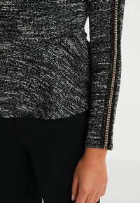 Top in maglia a trama bianco e nero con maniche lunghe e dettaglio in catena dorata sul lato, abbinato a pantaloni scuri.