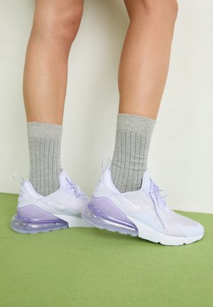 Ben iført grå ribstrikkede sokker og hvide Nike Air Max 270-sneakers med lilla luftkuddser, stående på grøn gulv.