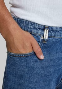 Jeans de mezclilla azul con un corte clásico, costuras visibles y un bolsillo delantero donde hay una mano. La etiqueta muestra "52 Studios".