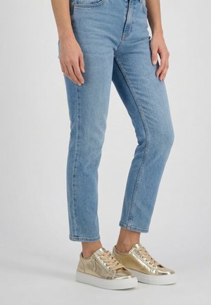 Person iført lyseblå jeans og metalliske guld sneakers med hvide såler, stående mod en ensartet lys baggrund.