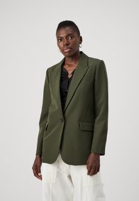 Blazer verde oliva con vestibilità sartoriale, chiusura a un bottone, revers a scollo e due tasche frontali, abbinato a un top nero.