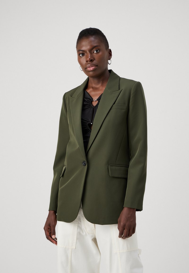 Blazer verde oliva con vestibilità sartoriale, chiusura a un bottone, revers a scollo e due tasche frontali, abbinato a un top nero.