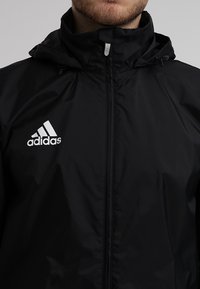 Mies, joka käyttää mustaa Adidas-hupparia, näkyvissä ylävartalo ja alahuuli.