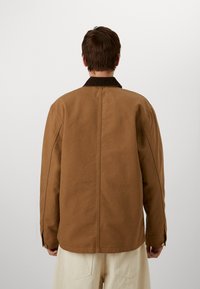 Giacca di cotone marrone con colletto in velluto a coste, caratterizzata da un taglio dritto, cuciture visibili e polsini con bottoni. Vista posteriore.