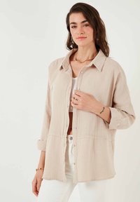 Chemise boutonnière beige clair en tissu texturé avec une coupe décontractée, dotée d'un col, de manches longues et de poignets retroussés.