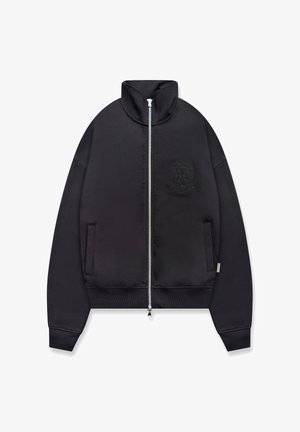 Svart jakke med hel glidelås, høy krage, ribbestrikkede mansjetter og nederkant, to frontlommer og et subtilt preget våpenskjold på brystet.