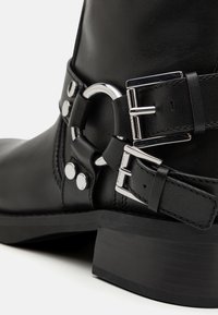 Bottine en cuir noir avec quincaillerie argentée, sangle à double boucle et finitions brillantes. Le talon est robuste et plat.