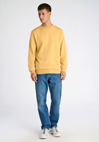 Felpa gialla con scollo rotondo e polsini a costine, abbinata a jeans di denim blu e sneaker bianche con dettagli multicolori.