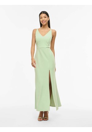 Žena stojaca a usmievajúca sa, oblečená v svetlozelených šatách bez rukávov s predným rozparkom a zlatými sandálmi s otvorenou špičkou na bielom pozadí.