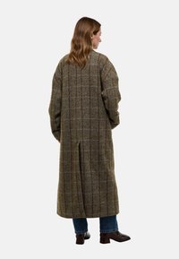 Long manteau en laine à motif écossais brun et vert, avec une coupe ample, des épaules tombantes et une fente au centre du dos. Porté sur un jean bleu.