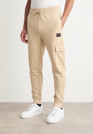 Joggers beiges en tissu doux, dotés d'une taille à cordon de serrage et d'une poche cargo sur le côté. Associés à des baskets blanches.