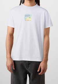 Valkoinen puuvillainen t-paita, jossa on värikäs kuva mehupaketista ja teksti "Cleptomanicx". Lyhyet hihat ja rento istuvuus.