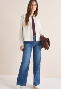 Camicia chiara con bottoni e due tasche sul petto, abbinata a jeans a gamba larga blu e sneakers bianche. Tiene in mano una giacca marrone.