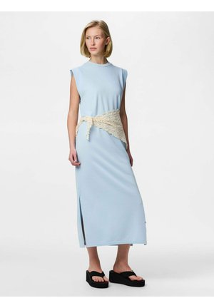 Žena oblečená v svetlomodrých midi šatách bez rukávov s bočnými rozparkami, béžovým čipkovým opaskom a čiernymi sandálmi na platforme, na bielom pozadí.