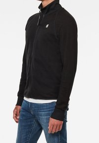 Svart zip-up jacka med hög krage, tillverkad av mjukt material. Har ribbade ärmavslut och en liten logotyp på vänster bröst. Bärs tillsammans med blå jeans.