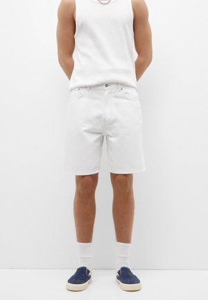 Weiße Denim-Shorts mit lockerer Passform, Fünf-Taschen-Design und dezenten Nähten. Kombiniert mit einem weißen Tanktop und marineblauen Slippers.