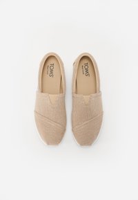 Zapatos beige tipo slip-on con una parte superior de tela texturizada, que cuentan con una abertura elástica en forma de V y suela de goma blanca. Logo de TOMS visible en el interior.