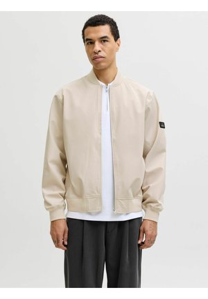 Ung mann iført en lys beige glidelåsjakke over en hvit skjorte og mørke løse bukser, stående mot en ensfarget bakgrunn.