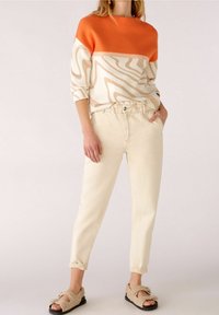 Maglione con righe orizzontali arancioni e beige, caratterizzato da un motivo a onde. Abbinato a pantaloni beige chiaro e sandali beige texturizzati.