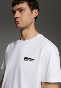 Biała bawełniana koszulka z czarnym logo na lewej piersi. Klasyczny okrągły dekolt i krótkie rękawy. Gładka tekstura i regularny krój.