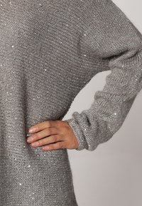 Harmaa neulepaita, jossa on löysä istuvuus, hopeiset koristekivet, leveät hihat ja teksturoitu kangas, korostaen rentoa muotoilua.