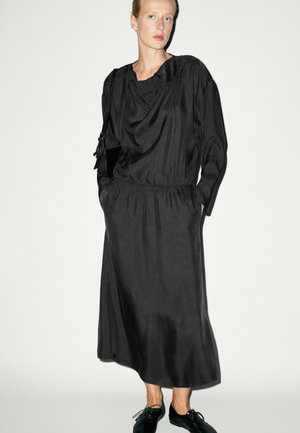 Frau trägt ein langes schwarzes Satinkleid mit langen Ärmeln, Hände in den Taschen, schwarze Tasche auf der Schulter und schwarze Schnürschuhe.