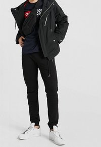 Black waterproof jacket with hood, layered over a navy graphic t-shirt, paired with black jogger pants and white sneakers.