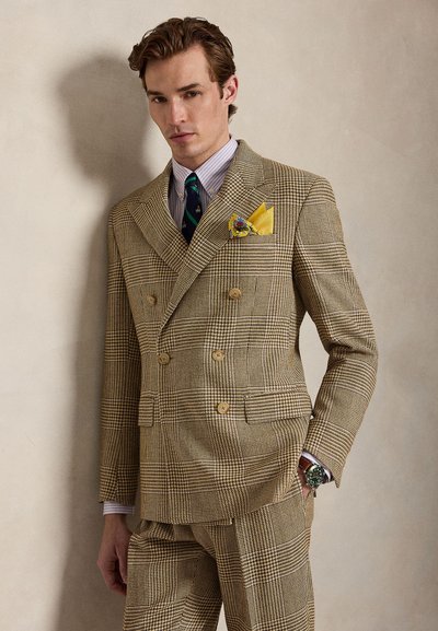 Muž v béžovom károvanom dvojradovom obleku s pásikavou košeľou, tmavozelenou kravatou, žltým vreckovkou a náramkovými hodinkami, stojaci oproti jednoliatej stene.