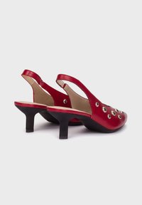Czerwone skórzane buty typu slingback z okrągłym noskiem, małym obcasem i dekoracyjnymi metalowymi ćwiekami na cholewce. Gładka faktura i kontrastująca czarna podeszwa.