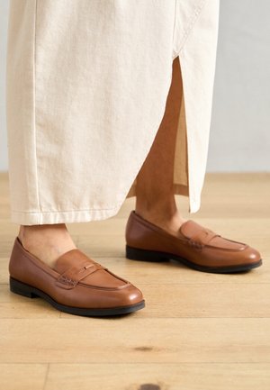 Lichtbeige rok met een zijsplit, gedragen met bruine leren loafers op een lichte houten vloer.