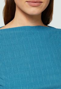 Haut texturé teal avec un design en tissu froissé, doté d'un grand col et d'une finition douce. Convient pour un look décontracté.