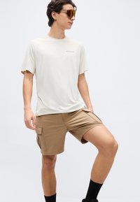 Νέος άνδρας που φοράει μπεζ cargo σορτς, ελαφρύ κρεμ t-shirt Patagonia, μαύρες κάλτσες και καθρεπτιστικά γυαλιά ηλίου, στέκεται με ένα γόνατο σηκωμένο.