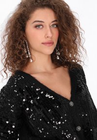 Zwarte pailletten cardigan met een diepe V-halslijn, twee juwelen knopen en een versierde ontwerp. Model met krullend haar en hangende oorbellen.