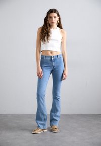 Lichtblauwe flared jeans met hoge taille en knoopsluiting, gecombineerd met een witte cropped haltertop en tan instappers. Soepele denimtextuur.