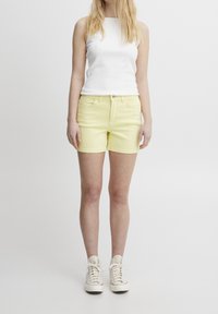 Keltainen farkkushortsit, joissa on kapea mitta, viisi taskua ja etunappi. Yhdistetty valkoisen hihattoman topin ja beigien tenkkareiden kanssa.