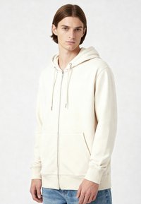 Felpa con zip di colore crema realizzata in tessuto morbido, dotata di cappuccio regolabile con cordino, ampie tasche frontali e polsini a costine.