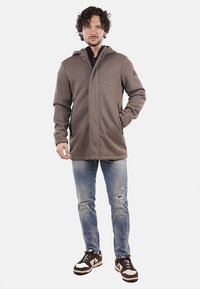 Giacca cappuccio marrone con una trama a costine, zip frontale e due tasche laterali, abbinata a jeans blu strappati e sneakers marroni.