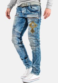 Jeans in denim celeste con ricami decorativi a forma di croce gialla e argentata, tasche con zip e design slim fit. Abbinati a sneakers bianche.